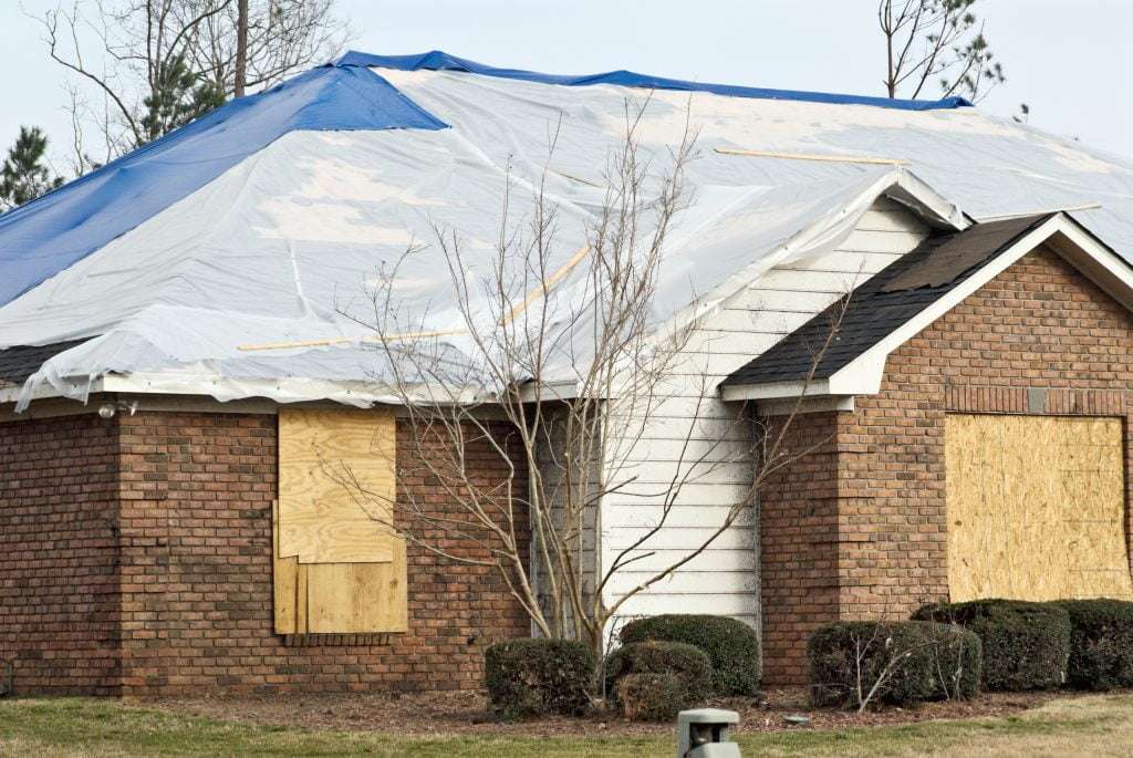 tornado damaged brick house with the windows boarded and a tarpaulin on the roof